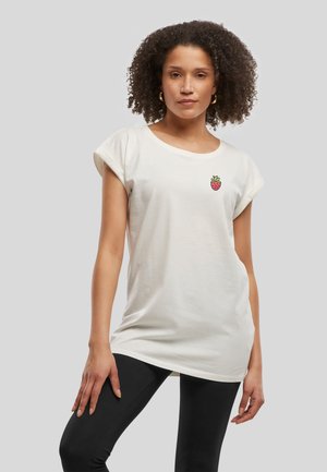 Femme aux cheveux bouclés portant un t-shirt blanc à manches courtes avec un petit écusson fraise et un legging noir, debout devant un fond gris.