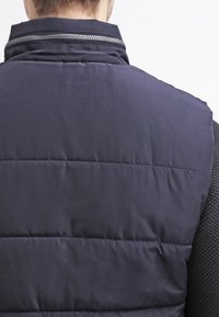 Granatowa kamizelka puchowa z poziomym pikowaniem, teksturowanym stójkowym kołnierzem i srebrnym zamkiem błyskawicznym. Gładki, syntetyczny materiał, odpowiedni do noszenia na warstwach.
