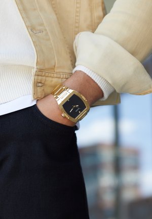 Pols met gouden metalen horloge met zwarte wijzerplaat, hand in de zak van een zwarte broek, beige jas met opgerolde mouwen en zichtbare witte trui.