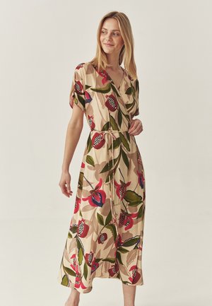 Woman wearing a beige wrap dress with red, green, and black floral patterns, tied at the waist, standing against a plain light background.