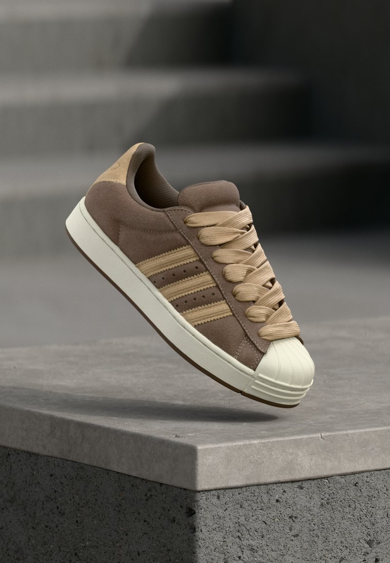 Zapatilla baja de ante marrón con acentos beige, puntera de goma color crema y cordones beige. Tres rayas paralelas en el lateral.