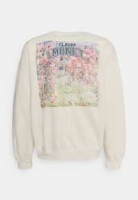 Krémová mikina s velkým grafickým potiskem na zádech zahrnujícím květinové umění a text "CLAUDE MONET." Má volný střih a žebrované manžety.