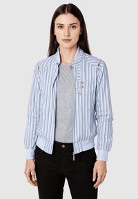 Joven mujer con cabello lacio castaño que lleva una chaqueta con cremallera a rayas azul claro y blanca sobre una camisa gris y pantalones negros, de frente.