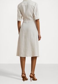 Ljusbeige linnklänning med skjortkrage, korta uppvikta ärmar och en utsvängd nederkant, i kombination med bruna högklackade sandaler.