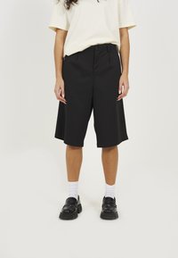 Black knee-length shorts with pleats, made of structured fabric. Worn with a light-colored t-shirt and black shoes, featuring chunky soles.