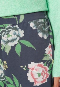 Ciemne spodnie w kwiatowy wzór z różowymi, białymi i zielonymi kwiatami, noszone z jasnoszarym swetrem widocznym na talii i nadgarstkach.