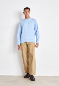 Sweatshirt bleu clair en tissu doux avec des poignets et un ourlet côtelés, arborant un petit logo noir. Associé à un pantalon beige ample et des bottes sombres.