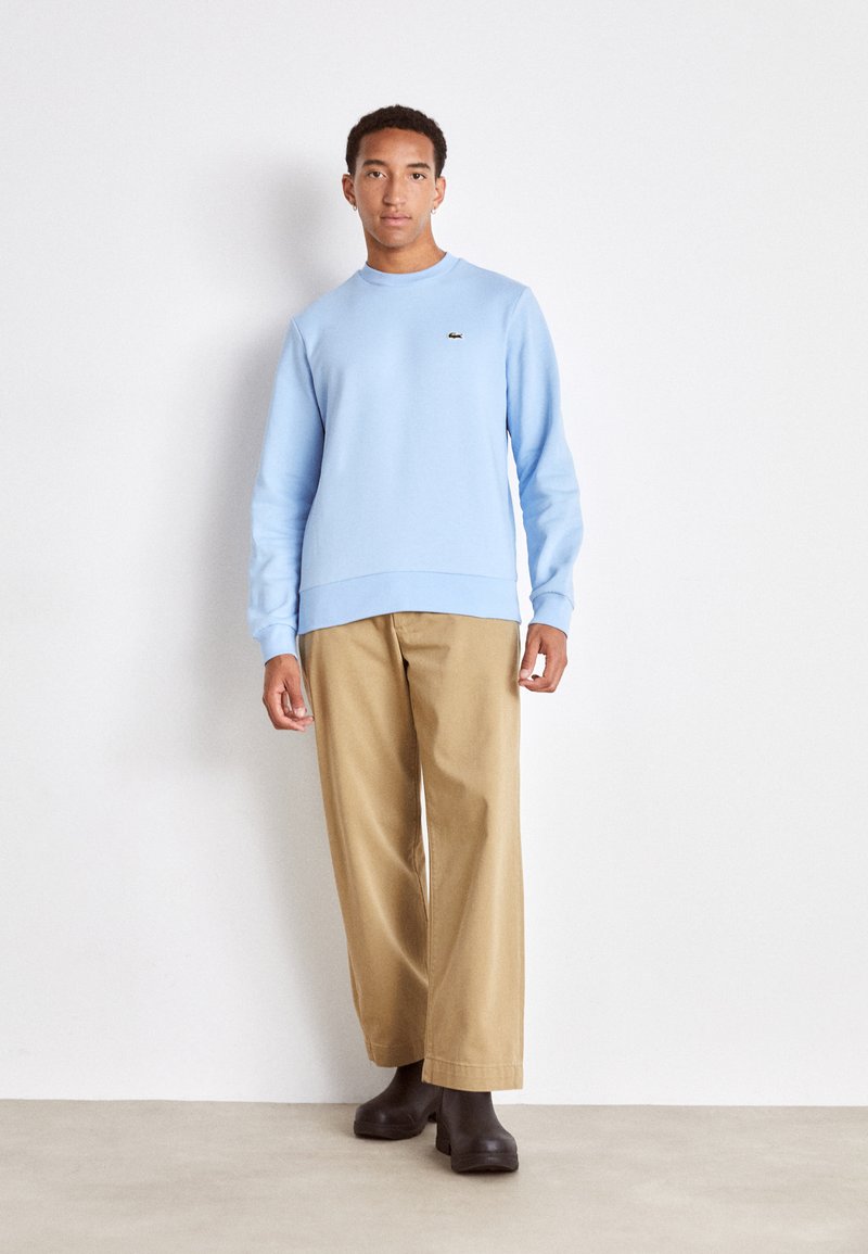 Lichtblauwe sweatshirt van zacht stof met geribbelde manchetten en zoom, voorzien van een klein zwart logo. Gecombineerd met losse beige broek en donkere laarzen.