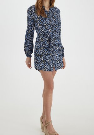 Nainen pukeutuneena mustaan, pitkähihainen kukkakuosiseen mekkoon, jossa on sinisiä ja valkoisia kukkia ja vyö, yhdistettynä beigenvärisiin kiilakorkoihin sandaaleihin.