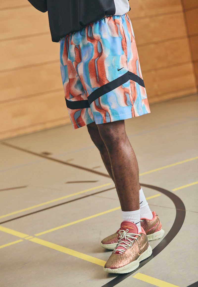 Nike rövidnadrág kék, piros és fehér absztrakt mintával. Fekete részletek az alján. Mettalik arany és zöld sportcipővel viselve.