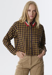 Camicia a quadri gialla e blu navy con colletto all'uncinetto che presenta strisce rosse, verdi e bianche. Parte anteriore con bottoni, maniche lunghe e vestibilità rilassata.