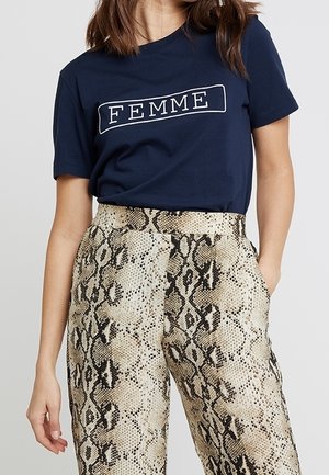 Kobieta ubrana w granatowy t-shirt z napisem „FEMME” oraz beżowe spodnie z wysokim stanem o wzorze imitującym skórę węża, z kieszeniami, stojąca z jedną ręką w kieszeni.