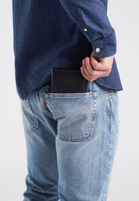 Svart läderplånbok delvis synlig i en bakficka på ljusblå jeans, tillsammans med en mörkblå mönstrad skjorta.