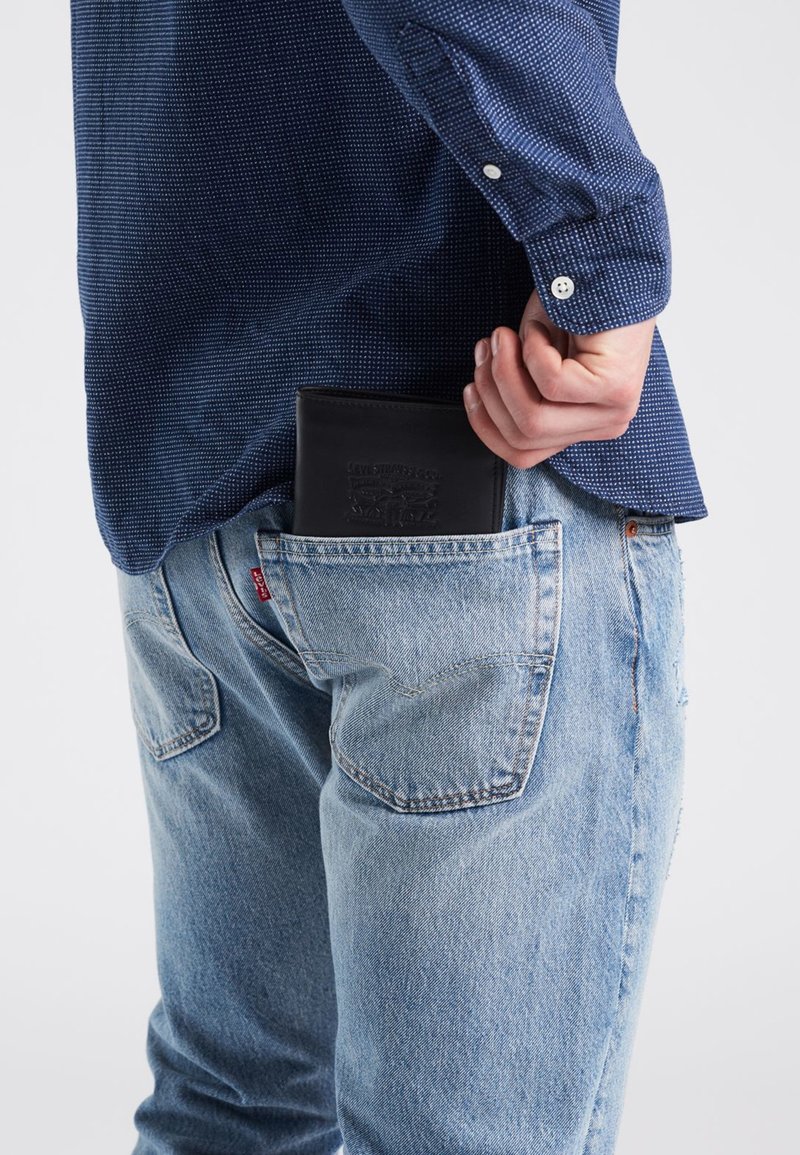 Svart läderplånbok delvis synlig i en bakficka på ljusblå jeans, tillsammans med en mörkblå mönstrad skjorta.