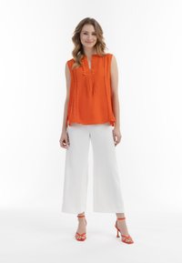 Orange ærmeløs top med broderede detaljer, parret med brede hvide bukser og orange sandaletter med stropper. Ensfarvet hvid baggrund.