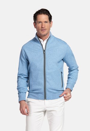 Homme portant un pull à fermeture éclair bleu clair avec des détails de fermeture éclair noirs et un pantalon blanc, debout devant un fond clair uni.