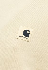 Μπαλώμα λογοτύπου Carhartt ραμμένο σε μπεζ υφή υφάσματος, που δείχνει το εμβληματικό στιλιζαρισμένο γράμμα "C" της μάρκας σε μαύρο χρώμα.