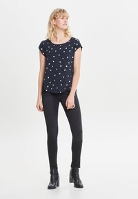 Marineblå top med guldprikker, korte ærmer og rund halsudskæring, parret med sorte skinny jeans og ankelstøvler. Glat stofstruktur.