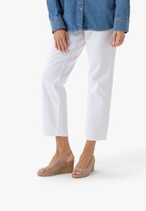 Mujer con pantalones blancos recortados, sandalias de cuña tostadas con punta abierta y camisa de mezclilla azul, de pie contra un fondo blanco.