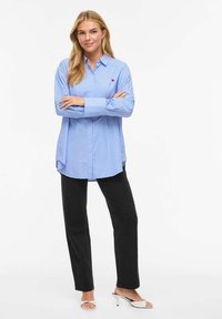 Chemise à rayures bleues et blanches avec boutons, manches longues, ourlet incurvé, et petit logo rouge sur la poitrine, associée à un pantalon noir.