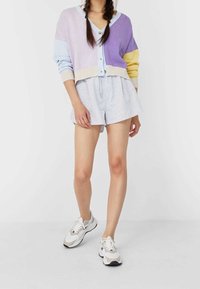 Pastelowy kardigan w odcieniach lawendy, niebieskiego i żółtego, połączony z jasnoszarymi szortami oraz białymi trampkami z beżowymi akcentami.