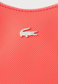 Rødt tekstureret stof med en glat, skinnende sølvkrokodille-logo. Designet har et subtilt mønster, der fremhæver holdbarhed og stil.