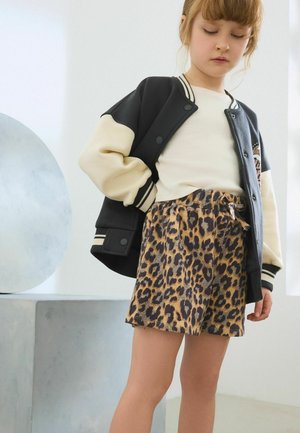 Ragazza giovane che indossa una giacca varsity nera e beige, top bianco e gonna con stampa leopardata, in piedi all'interno con un'espressione neutra.