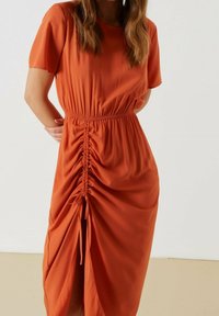 Šatové šaty v rezavě oranžové barvě s volným střihem, krátkými rukávy a nařaseným bočním švem s detaily na stažení pro nastavitelnou délku. Hladká textilní struktura.