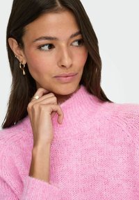 Roosa, kootud sviiter kõrge kaelusega ja tekstuuriga mustriga. Mudel toetab kätt lõuale, kandes kuldset ehtekunst ja minimaalset meiki.