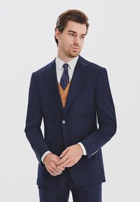 Granatowy garnitur o złożonym kroju, z pojedynczym guzikiem, klapami o kroju notched oraz dopasowanymi spodniami. Brązowa kamizelka i krawat w kropki dopełniają wygląd.