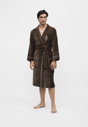 Homme debout pieds nus portant un peignoir marron foncé avec ceinture, deux poches avant et un logo blanc brodé sur la poitrine.