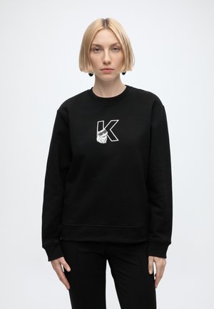 Giovane donna con capelli biondi corti indossa una felpa nera con una grande lettera bianca K e grafica di coniglio, in piedi su sfondo grigio uniforme.
