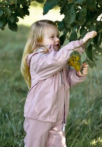 Vodoodporna svetlo roza jakna z elastičnimi manšetami, zadrgo in ujemajočimi hlačami. Otrok z dolgimi lasmi sega po zelenih listih, v ozadju pa je travnata površina.