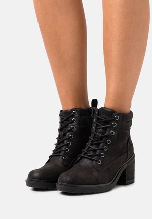Czarne sznurowane botki za kostkę na masywnych obcasach, noszone na gołych nogach, pokazane na jednolitym białym tle.
