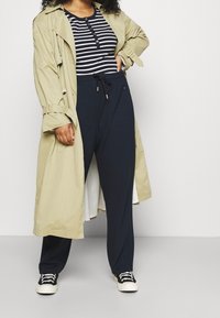 Beige trenchcoat over een marineblauwe gestreepte overhemd met knopen, gecombineerd met donkere joggingbroek en zwarte sneakers. Effen kleuren met heldere accenten.