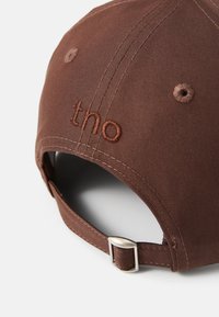Boné de beisebol castanho com textura de tecido. Apresenta um logótipo bordado "tno" e uma fivela de metal ajustável na parte de trás.