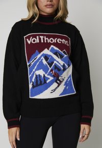 Donna che indossa un maglione nero con rifiniture rosse, caratterizzato da una toppa da sci di Val Thorens che mostra uno sciatore su montagne innevate blu.