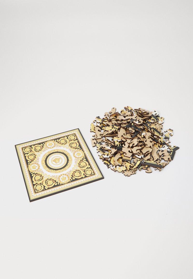 Square jigsaw puzzle with intricate gold and black design; pile of interlocking wooden pieces beside it. Textured surface with ornate patterns.