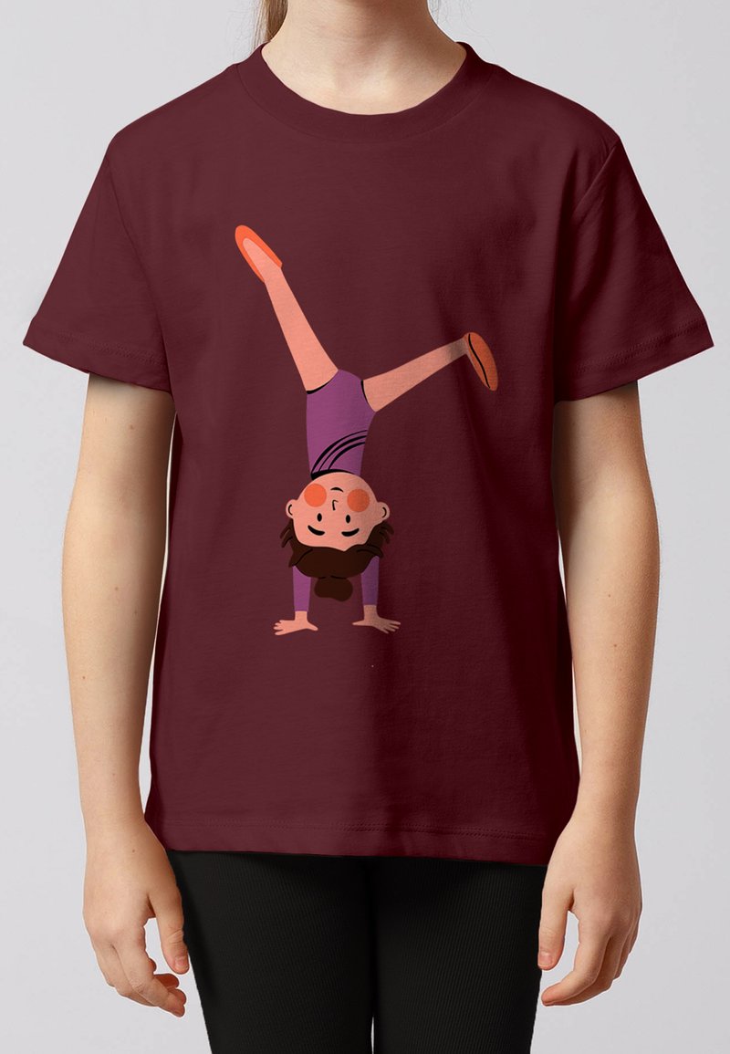 Enfant portant une chemise bordeaux avec un dessin animé d'une fille faisant un poirier en tenue violette et chaussures orange.