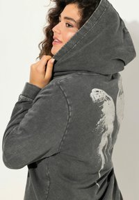 Sudadera gris con cordón, con un gran diseño abstracto blanco en la parte posterior. La tela parece suave con una textura ligeramente desgastada.