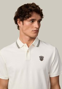 Biała koszulka polo z teksturowanej tkaniny, z czarno-białym paskiem przy kołnierzy oraz małym czarnym wyszywanym logo na piersi.