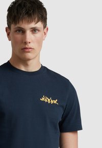 Granatowy bawełniany t-shirt z żółtym graficznym logo na lewej piersi. Krótkie rękawy i okrągły dekolt, gładka tekstura.