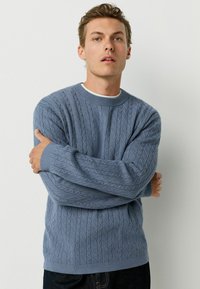 Light blue cable knit sweater with a crew neckline and ribbed cuffs, featuring a textured pattern and relaxed fit. Worn over a white shirt.