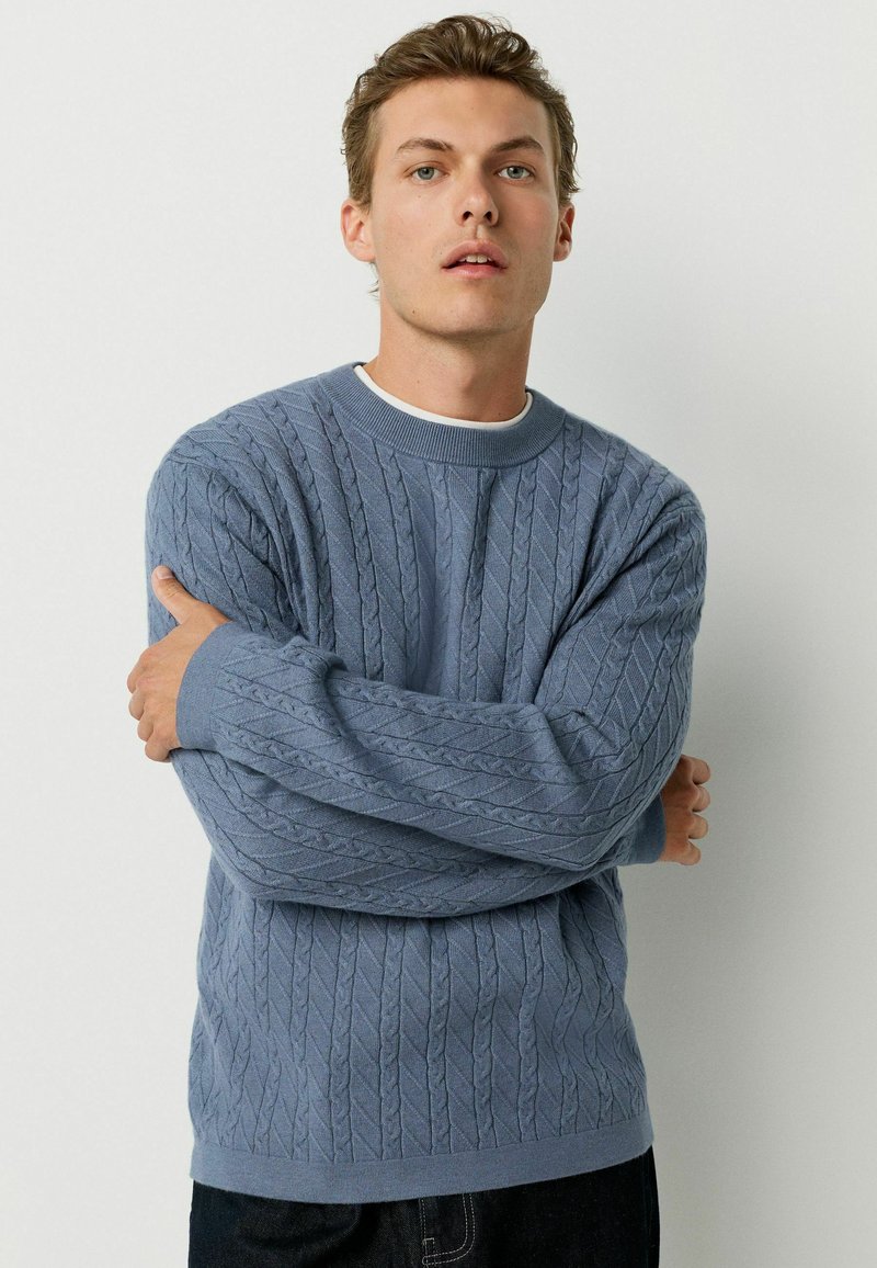 Light blue cable knit sweater with a crew neckline and ribbed cuffs, featuring a textured pattern and relaxed fit. Worn over a white shirt.
