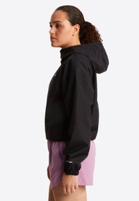 Γυναίκα με σγουρά μαλλιά δεμένα πίσω φορά μαύρο μπουφάν με κουκούλα και ροζ σορτς, όρθια πλάγια μπροστά σε λευκό φόντο.