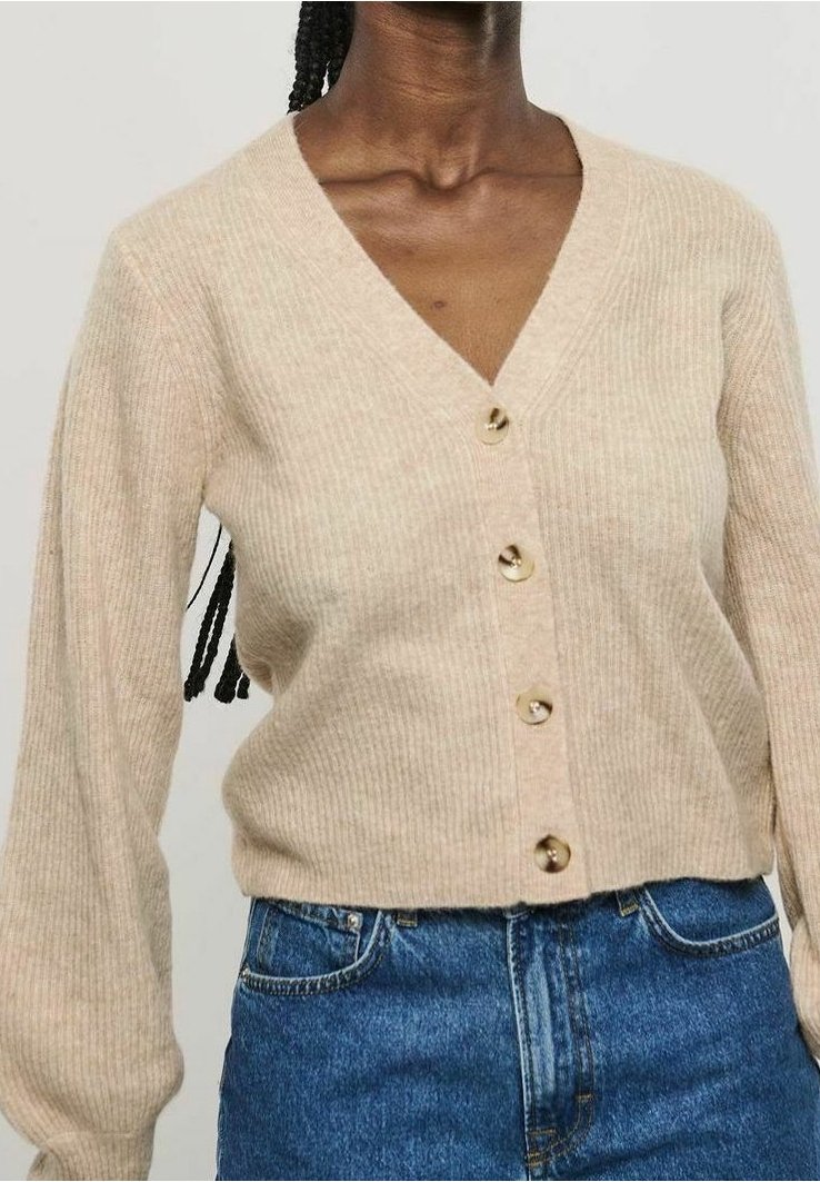 Porte un cardigan beige côtelé à col en V avec boutons et manches bouffantes, rentré dans un jean en denim bleu taille haute, raccourci au-dessus de la taille.