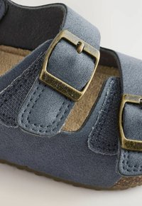 Modré semišové sandály se dvěma nastavitelnými zlatými sponami, texturovaným páskem a hnědým stélkou s jemným vzorem. Zaoblený design špičky.