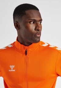 Oranje ritsjack met hoge kraag, met witte chevronstrepen op de mouwen en een logo op de borst. Glad, atletisch materiaal.