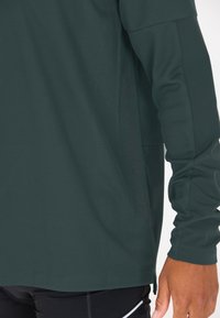 Dunkelgrünes Langarmshirt mit einem strukturierten Rautenmuster, seitlichen Schlitzen und einem dezenten Logo entlang der Naht. Weicher, elastischer Stoff.