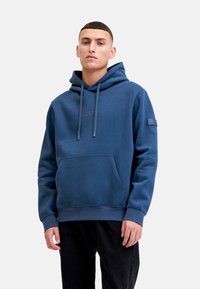 Joven vestido con una sudadera azul con capucha y bolsillo frontal, y pantalones negros, de pie frente a un fondo gris claro liso.